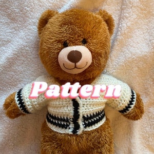 May include: A brown teddy bear wearing a cream-colored, crocheted jacket with black and white striped accents. The word "Pattern" is displayed across the jacket. The bear has a light brown snout and black eyes.