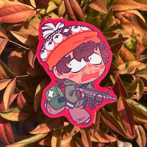 May include: A glittery sticker featuring a cartoon character with brown hair wearing an orange beanie with white patterns. The character has a wide-eyed, panicked expression, wears a green jacket, carries a green medical bag, and holds a rifle.