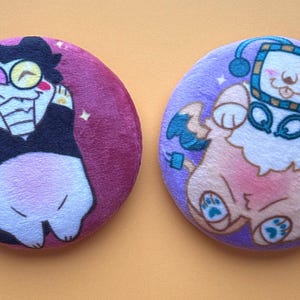 Puede incluir: Dos insignias redondas de felpa. Una es morada con un personaje de dibujos animados con traje negro y cuerpo blanco. La otra es morada con un personaje de perro de dibujos animados con auriculares. Ambos tienen una textura suave y afelpada.