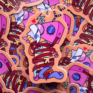 May include: A collection of colorful stickers featuring a cartoon robot-like character wrapped in red ribbon. The character has a pink head with blue eyes and a white face. The stickers have an orange background and a heart symbol.