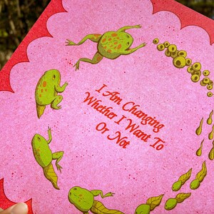 May include: A pink greeting card with a scalloped red border. The card features green frogs and tadpoles, with the text "I Am Changing Whether I Want To Or Not" in red.