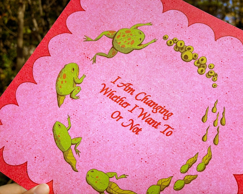May include: A pink greeting card with a scalloped red border. The card features green frogs and tadpoles, with the text "I Am Changing Whether I Want To Or Not" in red.