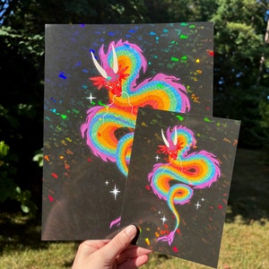 May include: Two prints of a rainbow dragon on a black background with iridescent glitter. The larger print is on the left and the smaller print is on the right.