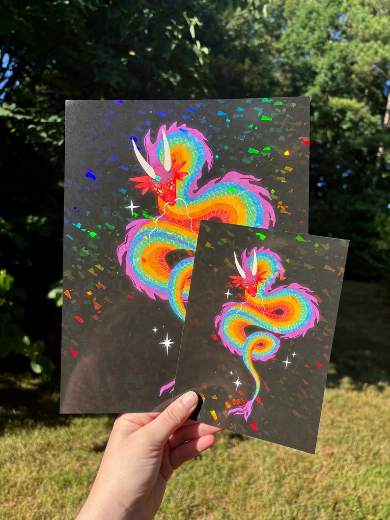 May include: Two prints of a rainbow dragon on a black background with iridescent glitter. The larger print is on the left and the smaller print is on the right.
