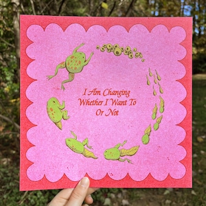 May include: A pink and red art print featuring a whimsical illustration of a frog's life cycle. The print includes the text "I Am Changing Whether I Want To Or Not" and is held by a hand.