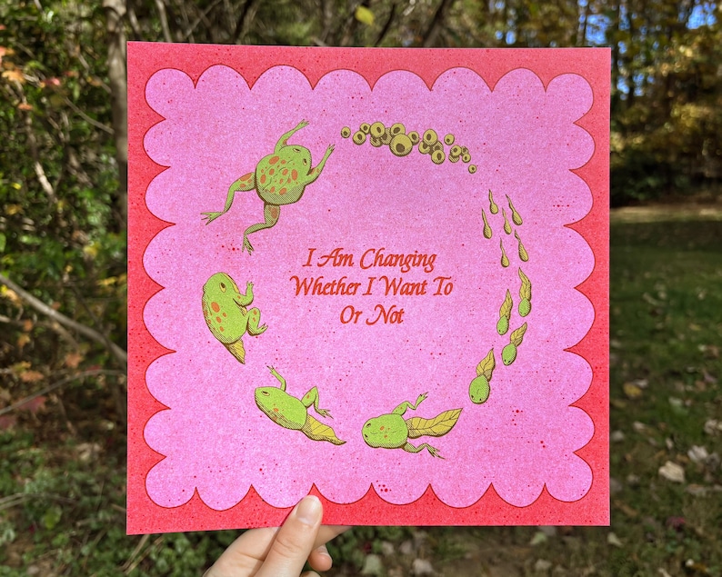 May include: A pink and red art print featuring a whimsical illustration of a frog's life cycle. The print includes the text "I Am Changing Whether I Want To Or Not" and is held by a hand.