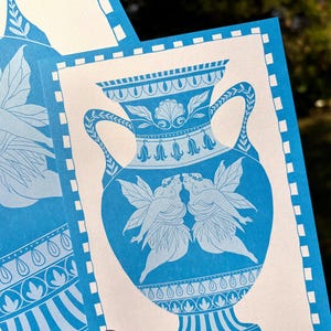 May include: Blue and white art print featuring a classical vase design. The vase is adorned with floral and shell motifs, and two figures with wings are kissing. The print has a border with a repeating pattern.