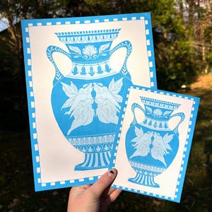 May include: Two blue and white art prints featuring a classical vase design with kissing fairies. The prints have a blue border and are held in a hand. The larger print is approximately 11x14 inches, and the smaller print is approximately 5x7 inches.