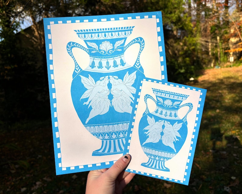 May include: Two blue and white art prints featuring a classical vase design with kissing fairies. The prints have a blue border and are held in a hand. The larger print is approximately 11x14 inches, and the smaller print is approximately 5x7 inches.