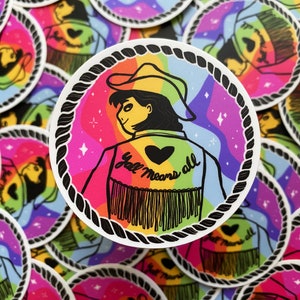 May include: A round sticker with a rainbow background and a cartoon illustration of a person wearing a cowboy hat and a denim jacket with the text "Y'all means all" on the back. The person is wearing a heart-shaped pin on their jacket.