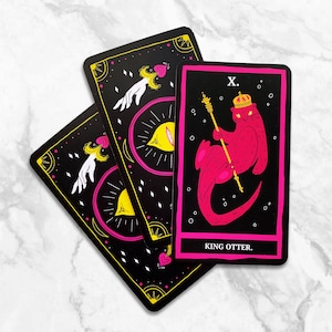 May include: A tarot card with a black background and yellow and pink accents. The card depicts a pink otter wearing a crown and holding a scepter. The card is labeled "King Otter" and "X."