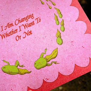 May include: Pink card with a scalloped edge featuring green frog illustrations and the text "I Am Changing Whether I Want To Or Not" in red. The card has a speckled texture.