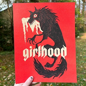 Peut inclure: Impression d'art rouge représentant une créature noire ressemblant à un loup, avec des dents et des griffes acérées. Le mot "girlhood" est écrit en police gothique. L'œuvre a un style illustratif vintage.