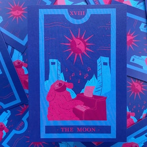 May include: A tarot card illustration featuring a pink creature playing a piano under a sun and moon design. The card is framed in blue with the text "THE MOON" at the bottom and Roman numerals at the top. Musical notes and architectural elements are also present.