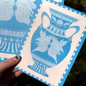 May include: Blue and white art print featuring a classical vase design with two figures kissing. The print has a decorative border and is held in a hand. The artwork is in a classical style.