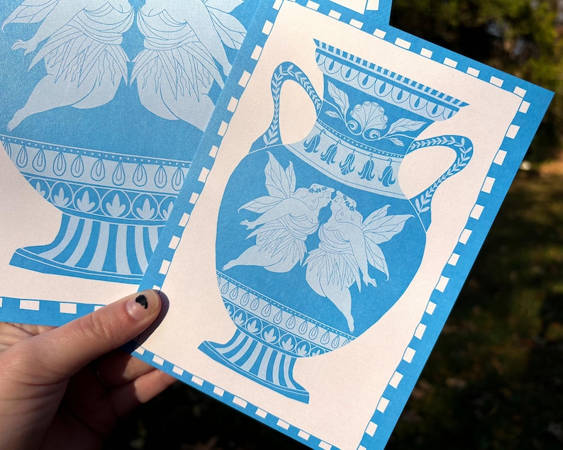 May include: Blue and white art print featuring a classical vase design with two figures kissing. The print has a decorative border and is held in a hand. The artwork is in a classical style.