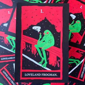 Puede incluir: Una pegatina negra y roja con una rana verde fumando un cigarrillo. La rana está sentada en una barandilla de madera. La pegatina dice "Loveland Frogman."