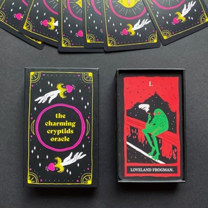 May include: A black box with the text "the charming cryptids oracle" on the front. The box contains a card with a green frog smoking a cigarette, sitting on a red bridge. The card has the text "I. LOVELAND FROGMAN." on it.