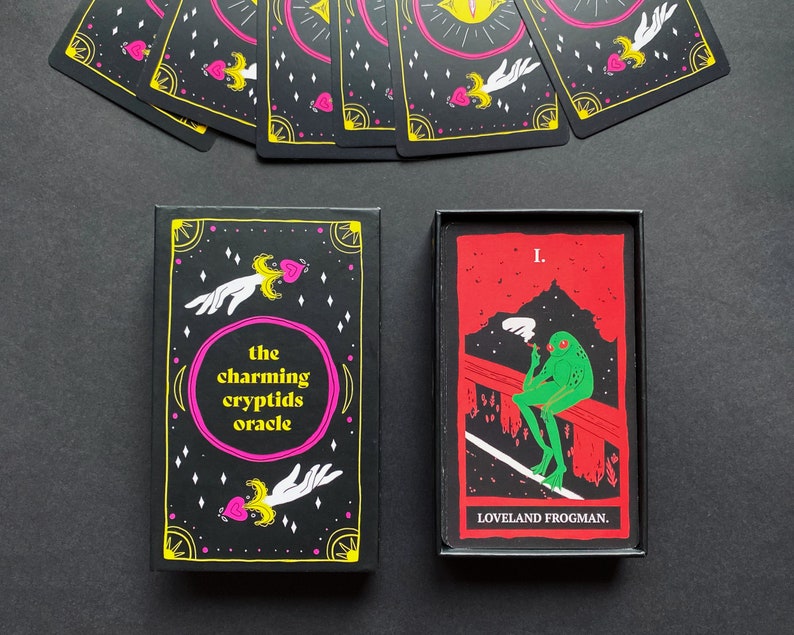 May include: A black box with the text "the charming cryptids oracle" on the front. The box contains a card with a green frog smoking a cigarette, sitting on a red bridge. The card has the text "I. LOVELAND FROGMAN." on it.