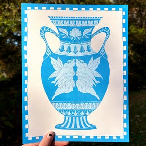 May include: A blue and white print of a decorative vase with handles, featuring a design of two fairies kissing. The vase has intricate floral and geometric patterns. The print is on a white background with a blue border.
