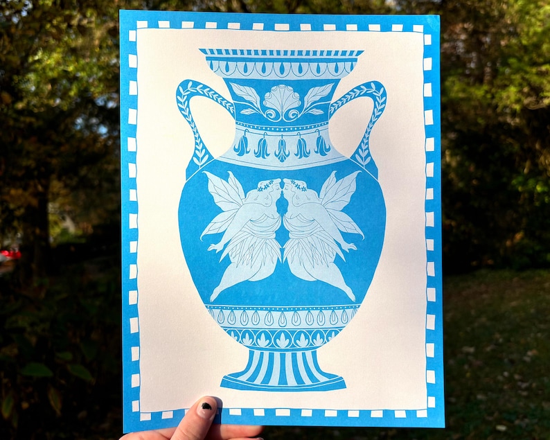 May include: A blue and white print of a decorative vase with handles, featuring a design of two fairies kissing. The vase has intricate floral and geometric patterns. The print is on a white background with a blue border.
