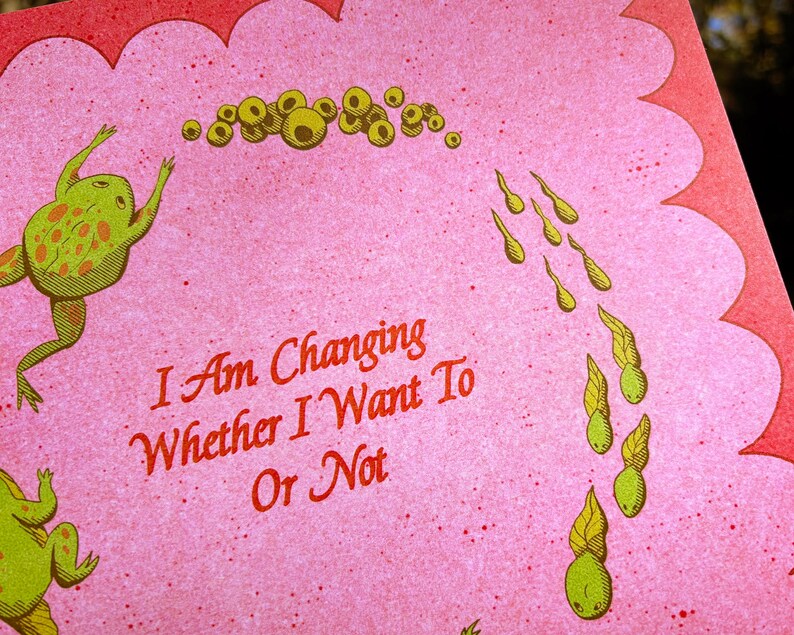 May include: A pink card with a scalloped edge features a whimsical illustration of frogs and tadpoles. The text "I Am Changing Whether I Want Or Not" is written in red script. The frogs are green with orange spots.
