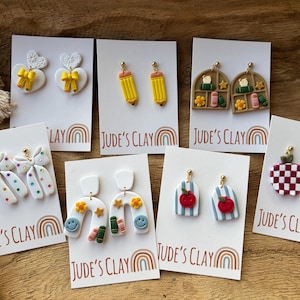 May include: Seven pairs of polymer clay earrings displayed on white cards labeled Jude's Clay. Designs include white hearts with yellow bows, yellow pencils, bookshelves, white bows with colorful dots, white arches with smiley faces, blue striped apples, and red checkered apples.