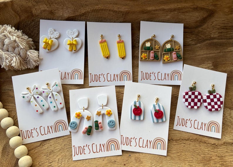May include: Seven pairs of polymer clay earrings displayed on white cards labeled Jude's Clay. Designs include white hearts with yellow bows, yellow pencils, bookshelves, white bows with colorful dots, white arches with smiley faces, blue striped apples, and red checkered apples.