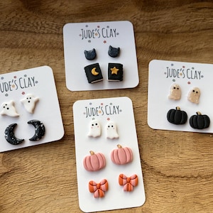 May include: Four white display cards labeled Jude's Clay featuring various stud earrings. Designs include white ghosts, black crescent moons, black cats, black books with gold stars and moons, beige ghosts, black pumpkins, pink pumpkins, and orange bows.