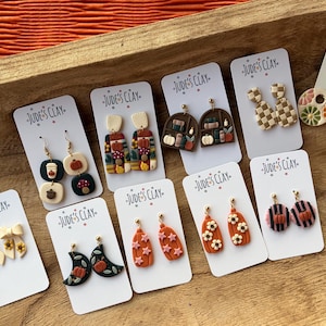 May include: A collection of ten pairs of polymer clay earrings displayed on white cards labeled Jude's Clay. Designs include pumpkins, mushrooms, coffee cups, moons, flowers, books, and checkered patterns in various autumn-inspired colors.