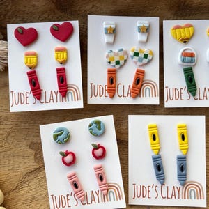 May include: Five white display cards labeled Jude's Clay featuring various school-themed polymer clay stud earrings, including shapes of red hearts, yellow pencils, colorful crayons, coffee cups, checkered hearts, books, globes, and red apples.