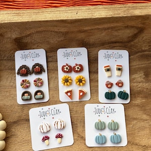 May include: Five white display cards labeled Jude's Clay, each featuring three pairs of small polymer clay stud earrings. Designs include brown sweaters, sunflowers, pumpkins, coffee cups, mushrooms, and pie slices in autumn-inspired colors.