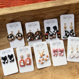 May include: Nine pairs of Jude's Clay earrings on white display cards. Designs include ghosts, pumpkins, black bows, and moons in various shapes like arches, diamonds, and bows. The earrings feature gold-toned hardware and colorful clay details.