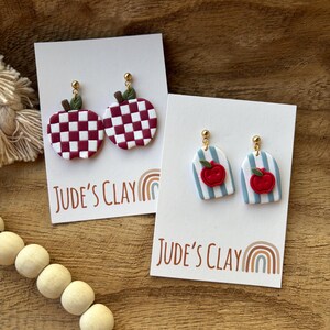 May include: Two pairs of dangle earrings on white display cards labeled Jude's Clay. One pair features maroon and white checkered apples with green leaves. The other pair features light blue and white striped arches with small red apple charms.