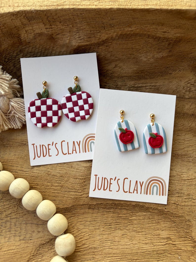 May include: Two pairs of dangle earrings on white display cards labeled Jude's Clay. One pair features maroon and white checkered apples with green leaves. The other pair features light blue and white striped arches with small red apple charms.