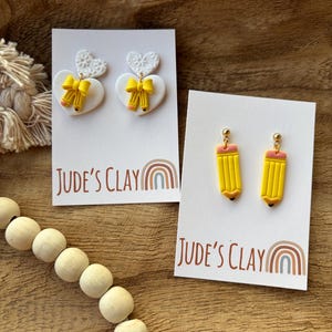 May include: Two pairs of earrings on white display cards labeled Jude's Clay. One pair features white heart-shaped studs with yellow pencil and bow charms. The second pair consists of yellow pencil-shaped drop earrings with pink erasers.