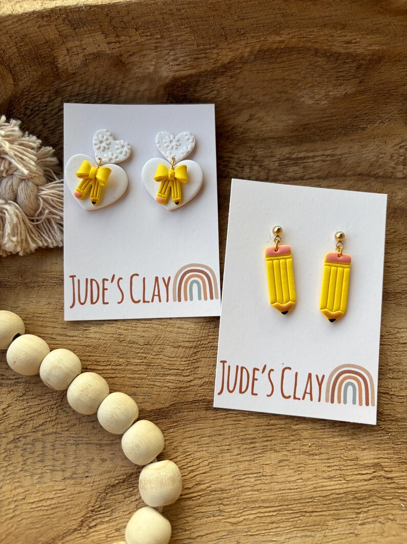 May include: Two pairs of earrings on white display cards labeled Jude's Clay. One pair features white heart-shaped studs with yellow pencil and bow charms. The second pair consists of yellow pencil-shaped drop earrings with pink erasers.