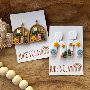 May include: Two pairs of polymer clay earrings on white display cards labeled Jude's Clay. One pair features tan arched windows with books, flowers, and stars. The other pair features white arches with smiley faces, flowers, stars, and books.