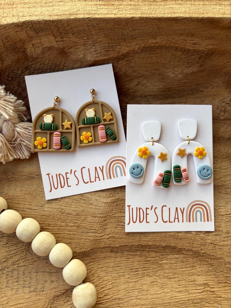 May include: Two pairs of polymer clay earrings on white display cards labeled Jude's Clay. One pair features tan arched windows with books, flowers, and stars. The other pair features white arches with smiley faces, flowers, stars, and books.