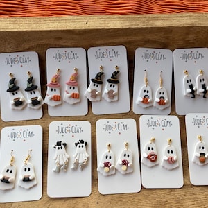 May include: Ten pairs of white ghost-shaped polymer clay earrings displayed on white cards labeled Jude's Clay. The ghosts feature various accessories including witch hats, pumpkins, mushrooms, coffee mugs, and black cats, all attached to gold-tone hooks.