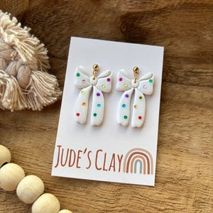 May include: A pair of white bow-shaped earrings featuring small, multicolored embedded rhinestones. The earrings are attached to a white display card with the text Jude's Clay and a rainbow graphic printed in brown and muted tones.
