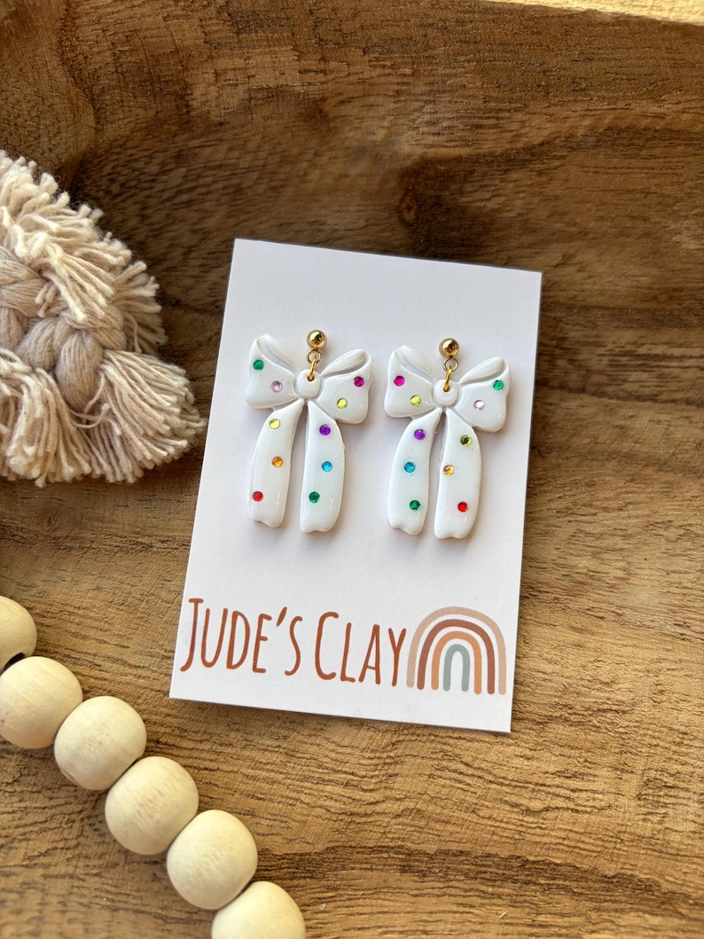 May include: A pair of white bow-shaped earrings featuring small, multicolored embedded rhinestones. The earrings are attached to a white display card with the text Jude's Clay and a rainbow graphic printed in brown and muted tones.