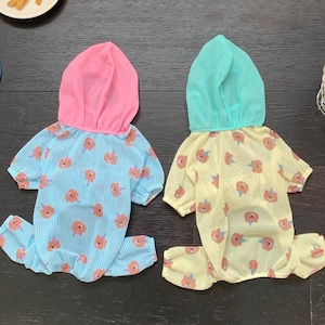 May include: Two pet jumpsuits. One is light blue with pink floral patterns and a pink hood. The other is yellow with pink floral patterns and a teal hood. Both have a striped pattern and are designed for small pets.