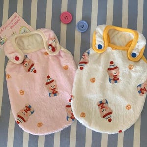 May include: Two plush dog vests, one pink and one white, with a repeating pattern of a bear wearing a hat and pretzels. The pink vest has a cream-colored collar and the white vest has a yellow collar. Pink and blue buttons are visible.
