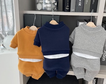May include: Three sets of pet clothing hang on wooden hangers. The sets include a sweatshirt and matching pants. The colors are mustard yellow, navy blue, and gray. Each set has a white undershirt peeking out from the bottom of the sweatshirt.
