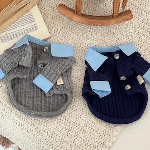 May include: Two dog sweaters, one gray and one navy blue, are displayed. Both sweaters have light blue collars and cuffs, and are buttoned. The sweaters are made of a ribbed knit material. The sweaters are designed to look like miniature blazers.