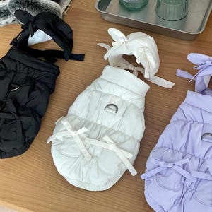 Könnte beinhalten: Eine Auswahl an gesteppten Haustierjacken in Schwarz, Weiß und Lavendel. Jeder Mantel hat eine Schleife am Hals und an der Taille, mit passendem Hut. Die Mäntel haben einen D-Ring zum Anbringen der Leine.