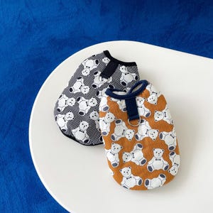 Peut inclure: Deux gilets pour chiens avec des motifs d'ours en peluche. L'un est noir et blanc à rayures verticales, l'autre est marron à rayures horizontales. Les deux ont une bordure bleu marine et une petite boucle pour une laisse.