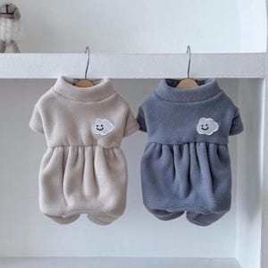 May include: Two fleece pet sweaters, one in beige and one in slate blue, each featuring a high collar, short sleeves, and a gathered skirt section. A small white embroidered cloud patch with a smiling face is centered on the chest of each.