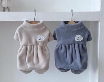 May include: Two fleece pet sweaters, one in beige and one in slate blue, each featuring a high collar, short sleeves, and a gathered skirt section. A small white embroidered cloud patch with a smiling face is centered on the chest of each.